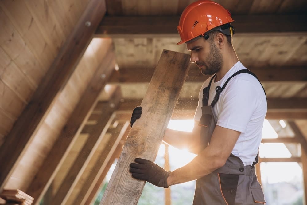 A construction worker wearing an orange safety helmet and gloves, inspecting a wooden beam inside a building under construction.