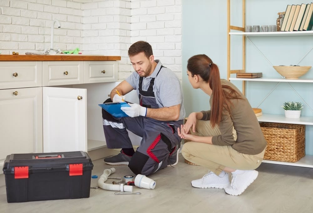 A technician inspecting plumbing pipes under a kitchen sink while a woman observes, in a modern kitchen setting.