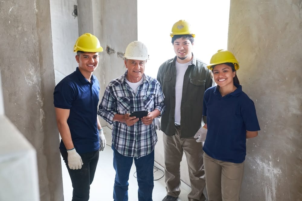 A construction team of four workers, including a supervisor with a tablet, standing inside a building under construction, wearing safety helmets and casual work attire.