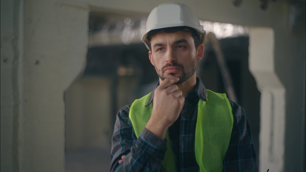 A construction worker in a hard hat and safety vest thoughtfully inspecting a building site under construction.