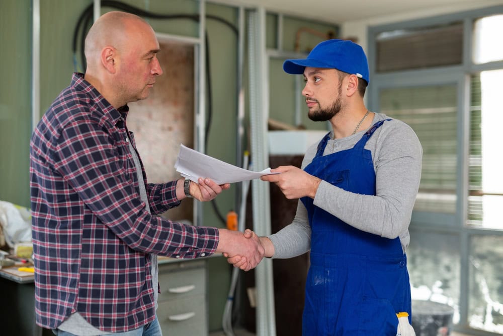 Two men shaking hands indoors, one in casual clothes and the other in a blue work uniform, symbolizing agreement or partnership in the construction or contracting industry.