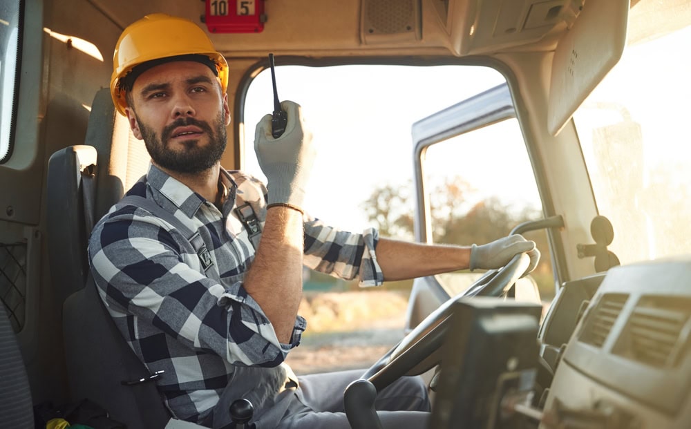 A construction worker wearing a yellow safety helmet and plaid shirt, sitting in the driver's seat of a construction vehicle, holding a walkie-talkie, with a sunny outdoor background.