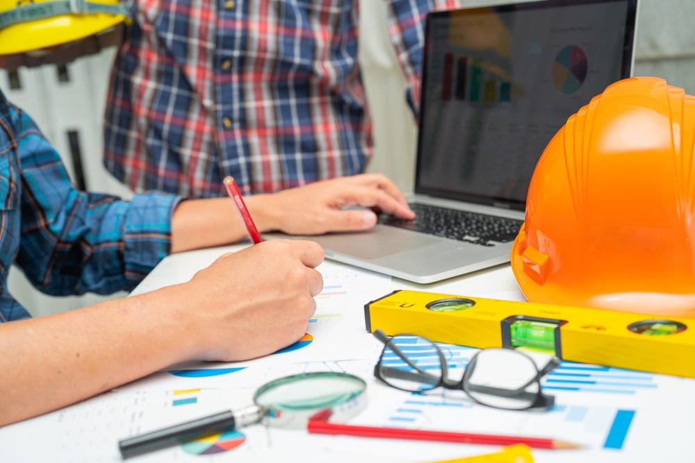A construction professional reviewing project plans with a laptop, safety helmet, and measuring tools on a desk, emphasizing planning and safety in the contracting industry.