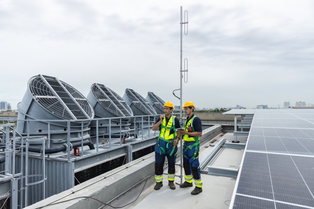 An industrial rooftop with two workers in safety gear inspecting large HVAC units and solar panels under a cloudy sky.