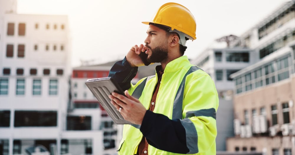 A construction worker wearing a yellow safety helmet and high-visibility vest, holding a tablet and talking on the phone at a building site with modern structures in the background.