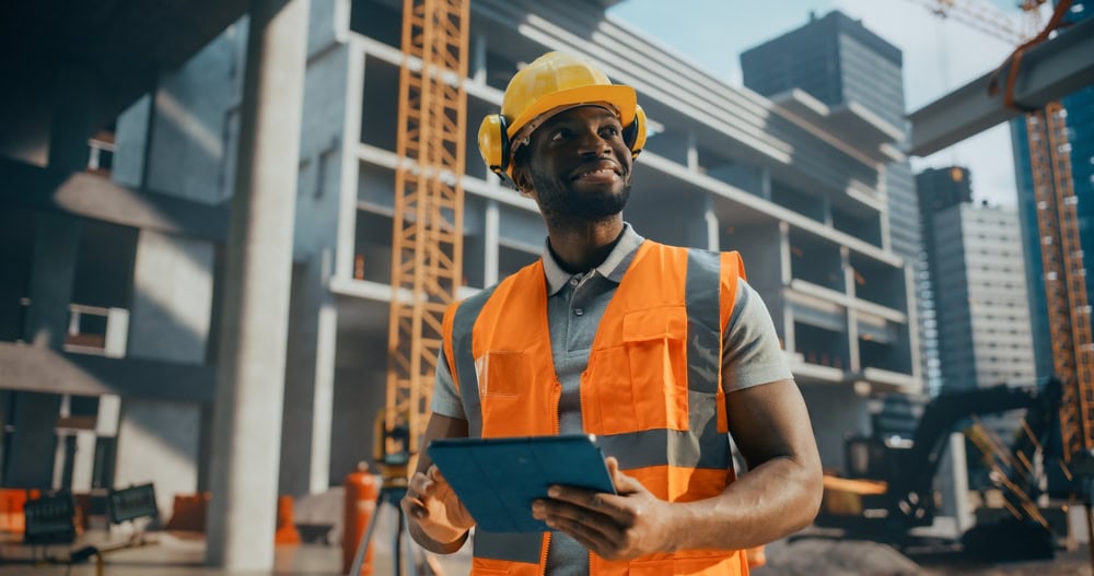 A construction worker wearing a yellow safety helmet and orange safety vest holding a tablet at a building site with cranes and partially constructed buildings in the background.