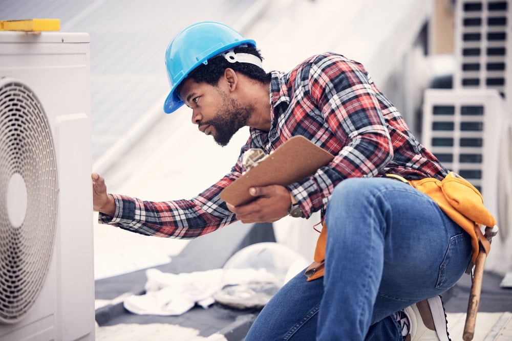 An HVAC technician inspecting an outdoor air conditioning unit, holding a clipboard, wearing a blue safety helmet and plaid shirt, on a rooftop with HVAC equipment in the background.