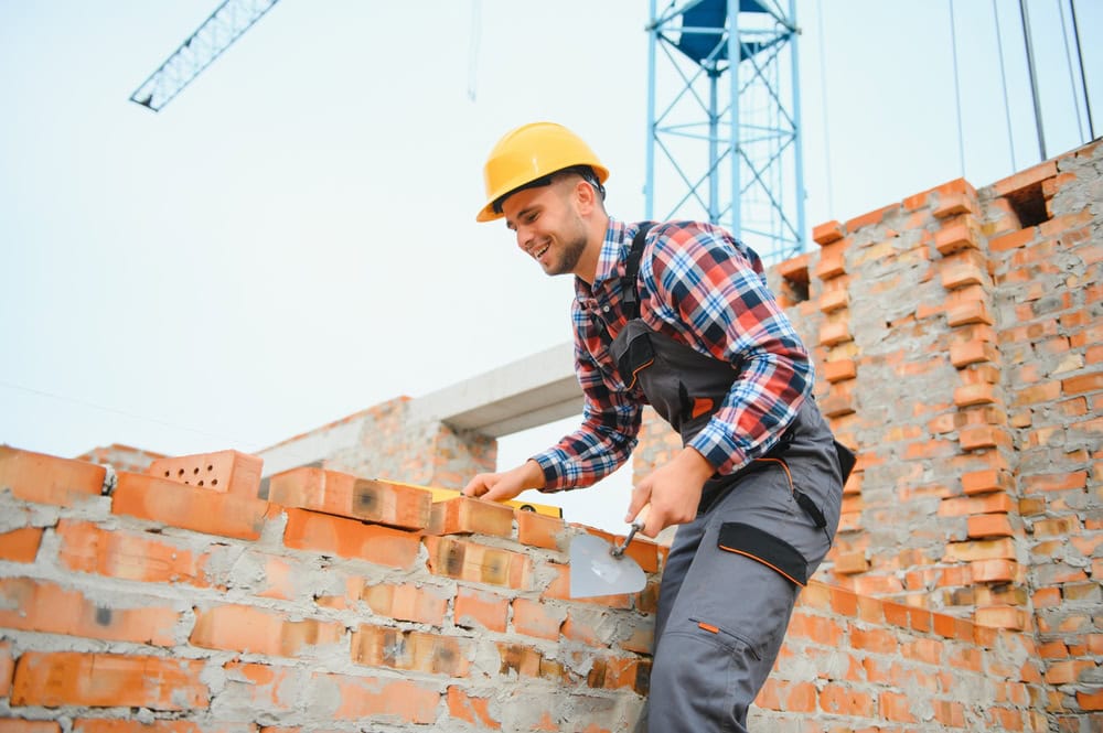 A construction worker wearing a yellow safety helmet and plaid shirt building a brick wall on a construction site with cranes in the background.