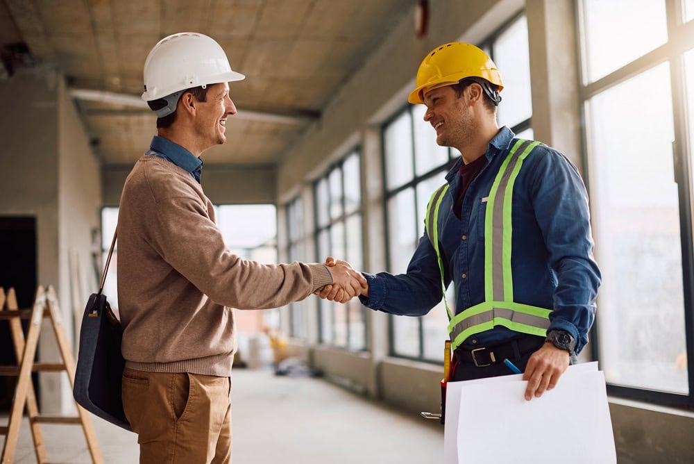 A construction professional and a worker in safety gear shaking hands inside a building under construction, symbolizing partnership and collaboration in the contracting industry.