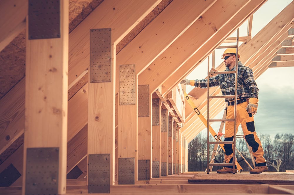 A construction worker inspecting wooden framing on a building site, wearing safety gear and using a level, with a clear sky and trees in the background.