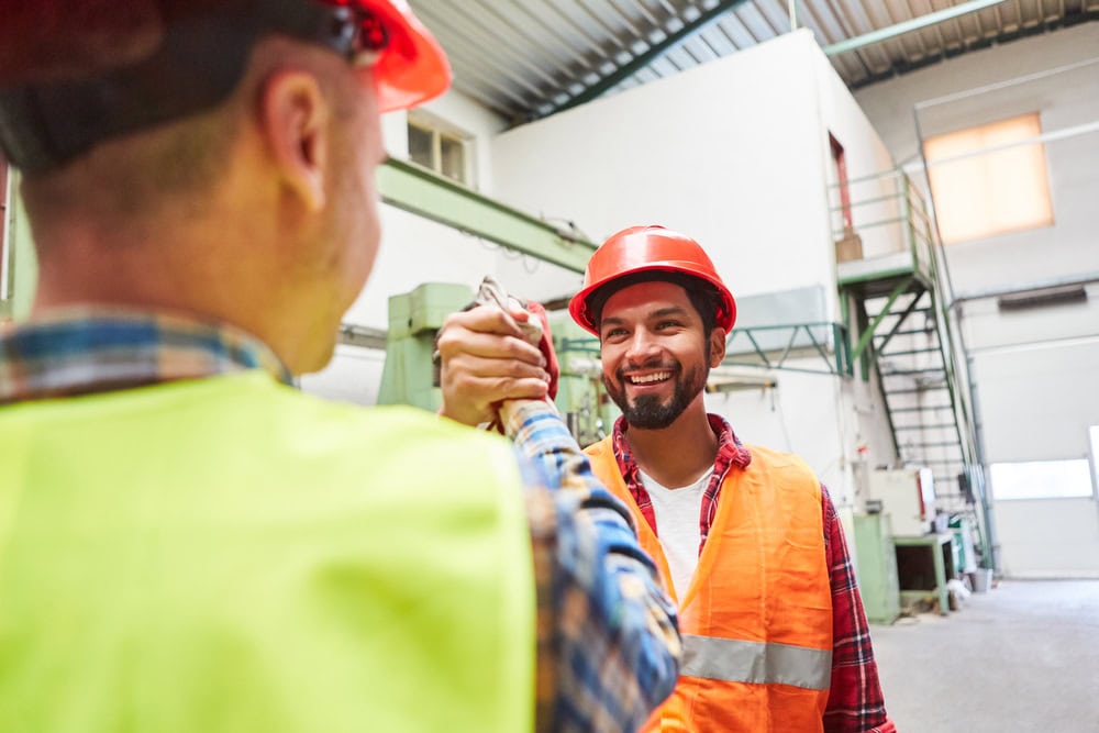 Construction workers shaking hands inside a warehouse, wearing safety gear including helmets and vests, symbolizing teamwork and professionalism in the contracting industry.