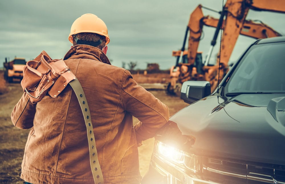 A construction worker inspecting a vehicle at a construction site with heavy machinery in the background.