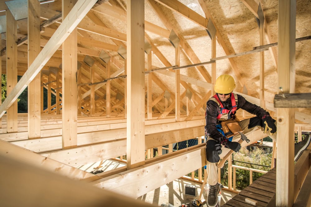 A construction worker wearing a yellow safety helmet and harness working on a wooden framework of a building under construction.