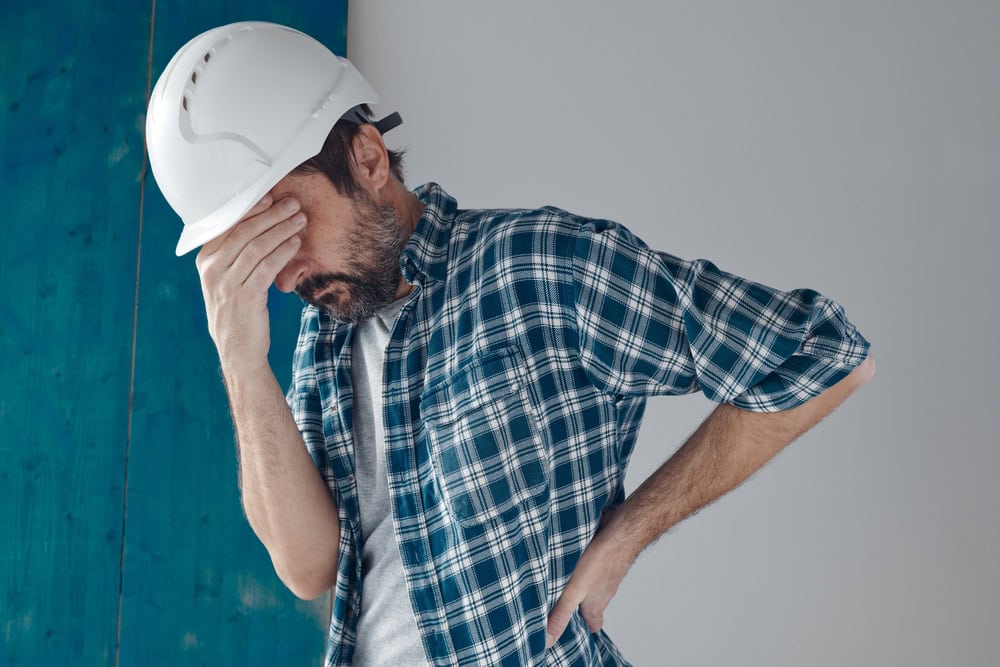 A man wearing a white construction helmet and plaid shirt appears stressed or frustrated, holding his forehead with one hand, standing against a plain background.