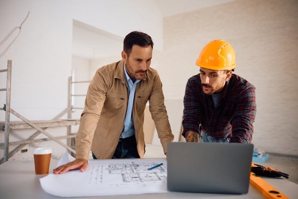 A construction project discussion between two men, one in a hard hat and the other in casual attire, reviewing blueprints and plans on a laptop in an unfinished building.