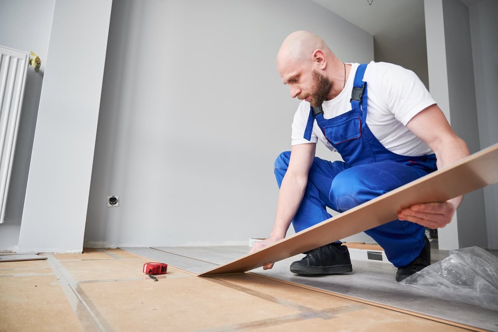 A construction worker installing flooring, kneeling on the floor with tools and materials in a modern room, preparing the surface for new flooring installation.