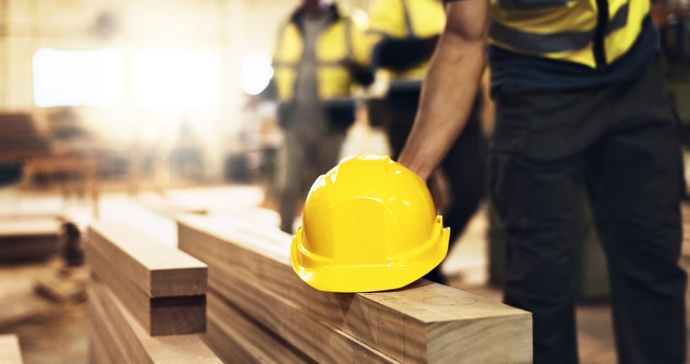 A yellow safety helmet resting on a wooden beam in a construction site with workers in the background, emphasizing safety and professionalism in the contracting industry.