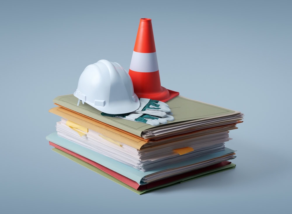 A construction safety helmet, traffic cone, and gloves placed on top of organized contractor documents and blueprints, symbolizing construction project planning and safety measures.