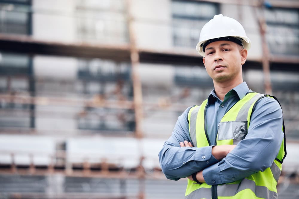 A construction site supervisor wearing a white safety helmet and a high-visibility vest, standing confidently with arms crossed in front of a building under construction.