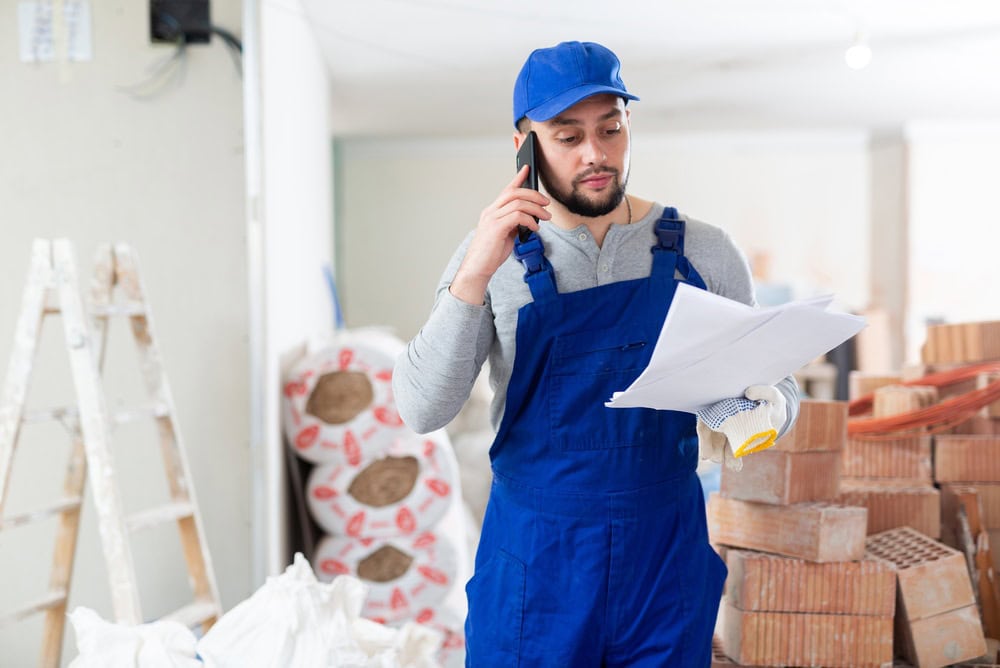 A construction worker in blue overalls and a cap talking on a mobile phone while reviewing documents at a building site with bricks and supplies in the background.