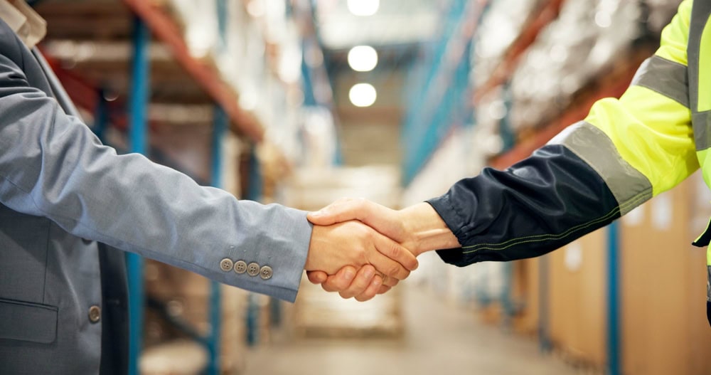 Business professionals shaking hands in a warehouse, symbolizing partnership and collaboration in the construction or contracting industry.