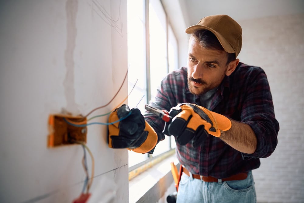 Electrician installing wiring in a wall, wearing safety gloves and a brown cap, working on electrical outlet at a construction site.