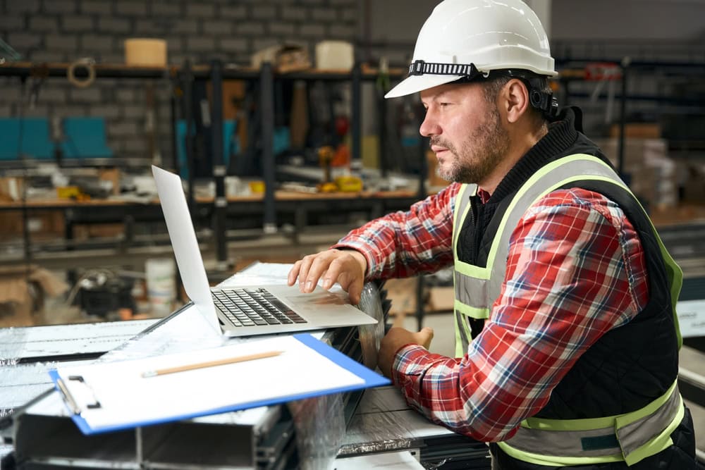 A construction worker wearing a white safety helmet and reflective vest working on a laptop in a workshop or construction site.