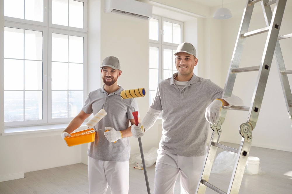 A smiling handyman holding a paint roller and a spray paint gun, standing in a bright, empty room with large windows, preparing for painting or renovation work.