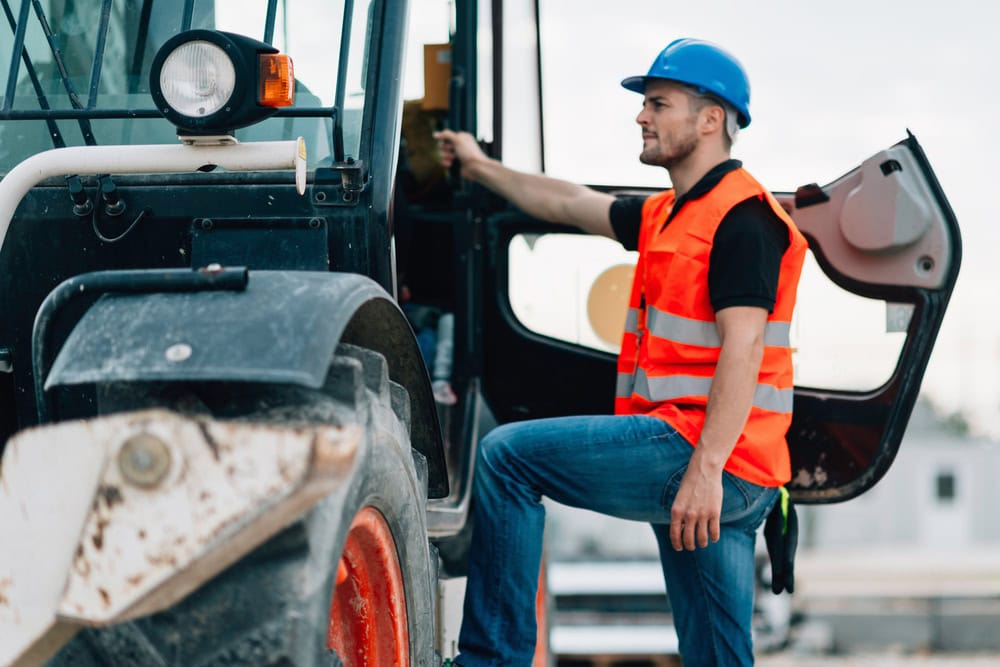 Construction worker in a blue helmet and orange safety vest sitting on a tractor, preparing for construction work, emphasizing contractor licensing and safety.