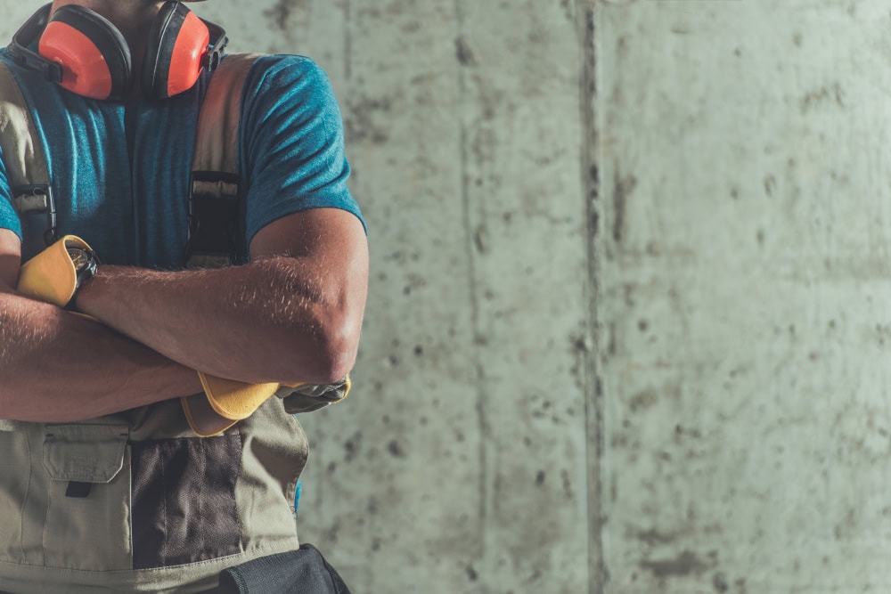 Construction worker wearing safety gear, including headphones and gloves, standing against a concrete wall, emphasizing construction safety and licensing.