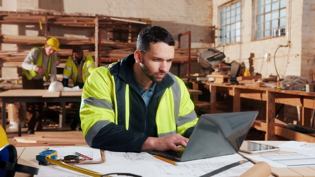 Construction professional working on a laptop in a workshop with other workers in the background, emphasizing contractor training and licensing.