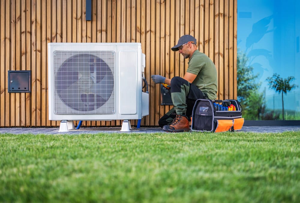 HVAC technician servicing an outdoor air conditioning unit with tools and a toolbox on a grassy lawn near a wooden wall.