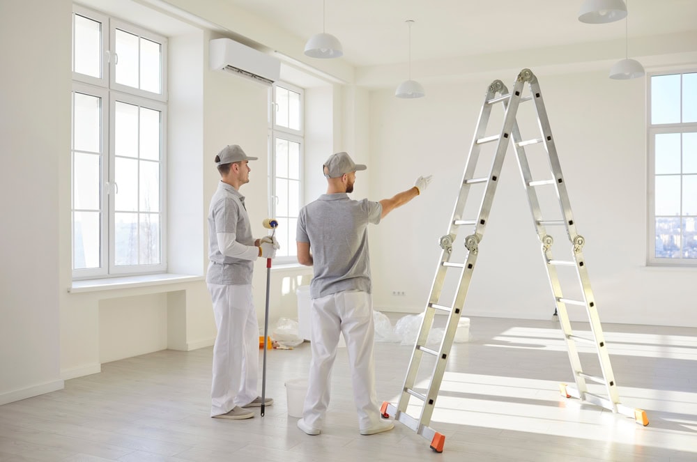Two professional contractors inspecting a bright, spacious room with large windows, a ladder, and painting tools, preparing for renovation or painting work.