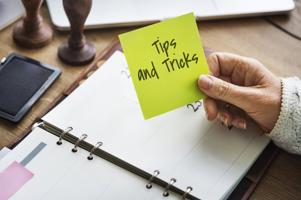 Hand holding a yellow sticky note with 'Tips and Tricks' written on it, placed over a planner on a wooden desk with office supplies.