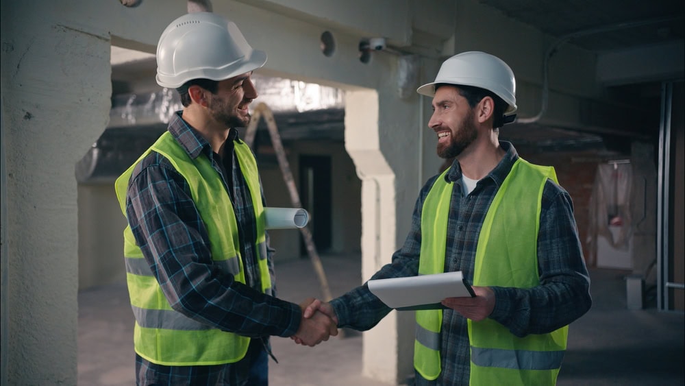 Two construction professionals in safety vests and helmets shake hands on a building site, discussing project plans for contractor licensing.