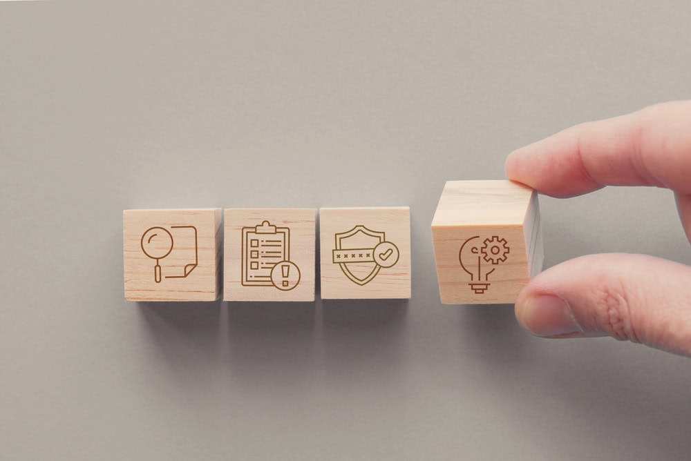 Hand placing a wooden block with a gear icon next to other blocks with icons representing tools, documents, and safety, symbolizing contractor licensing and certification.