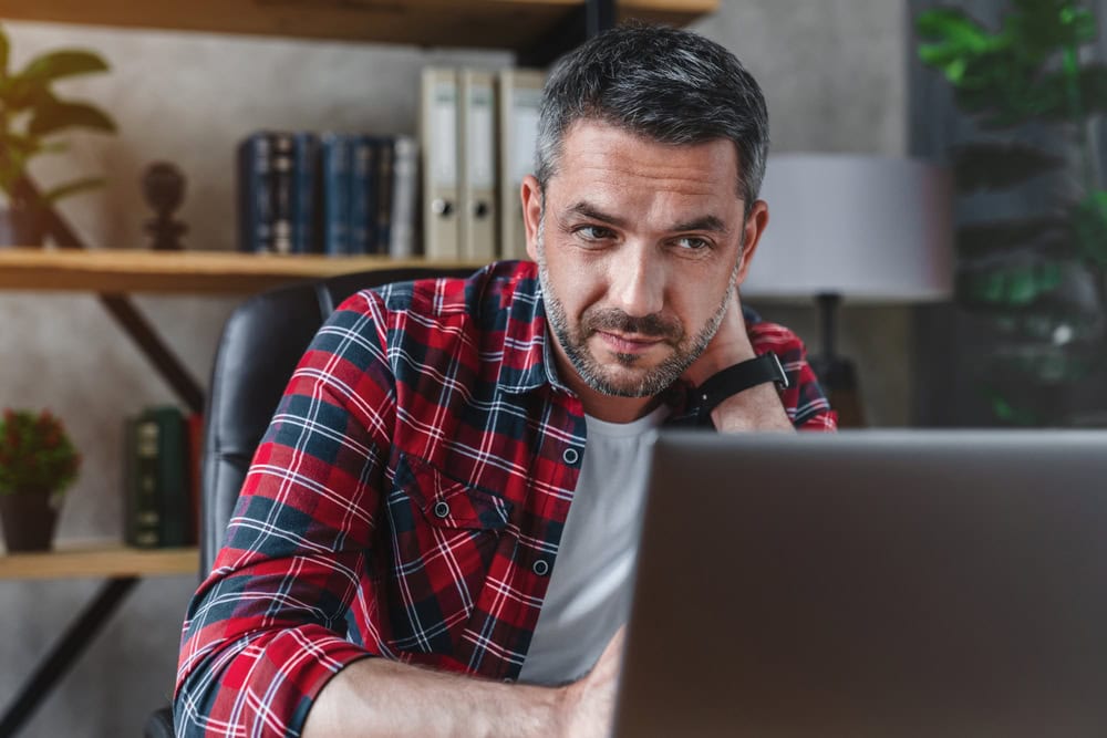 Man working on a laptop in a modern office setting, focused and thoughtful, with books and plants in the background, emphasizing professional contractor training and licensing.