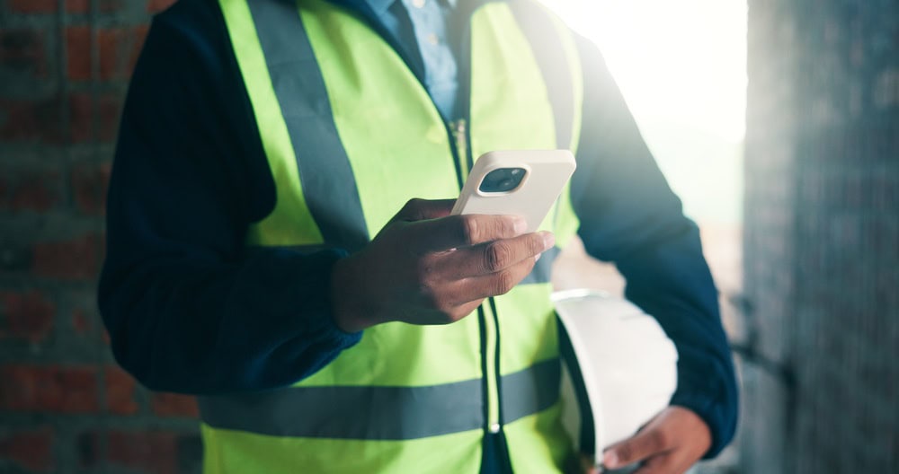Construction worker in a high-visibility vest using a smartphone at a construction site.