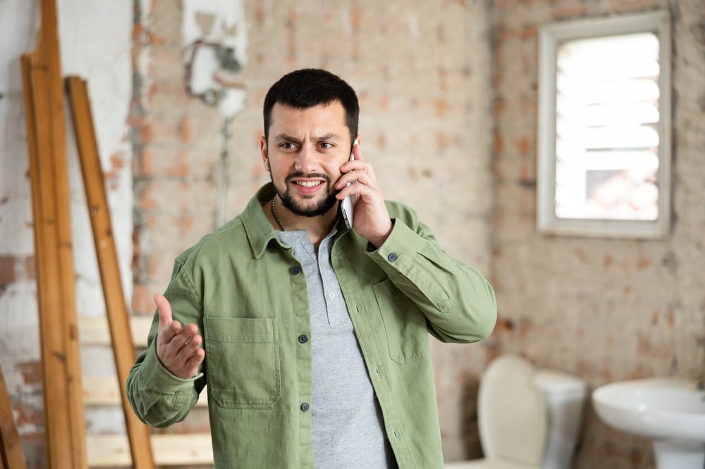 Man in green jacket talking on phone inside a construction or renovation site with exposed brick walls and a window.