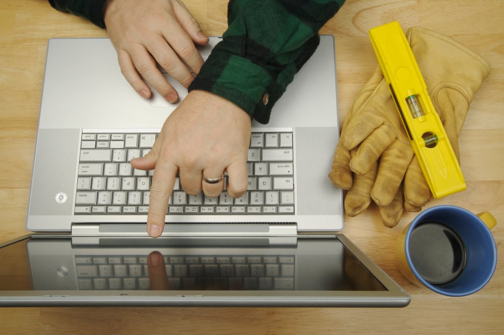 Person using a laptop with construction gloves, level, and coffee mug on a wooden desk, illustrating contractor licensing and online learning.